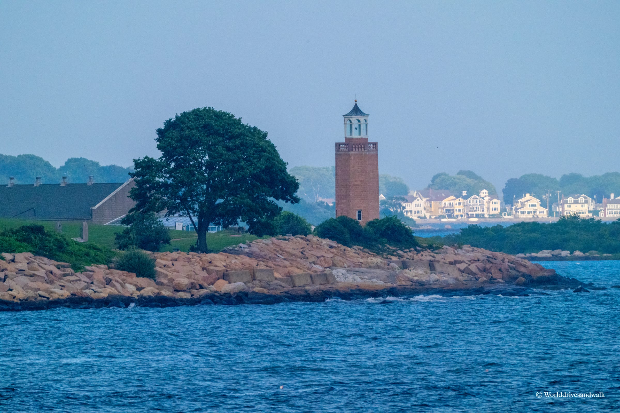 A red brick lighthouse with a white cupola stands on a rocky point across blue water, with a large shade tree and residential buildings visible along the distant shoreline under clear sky.
