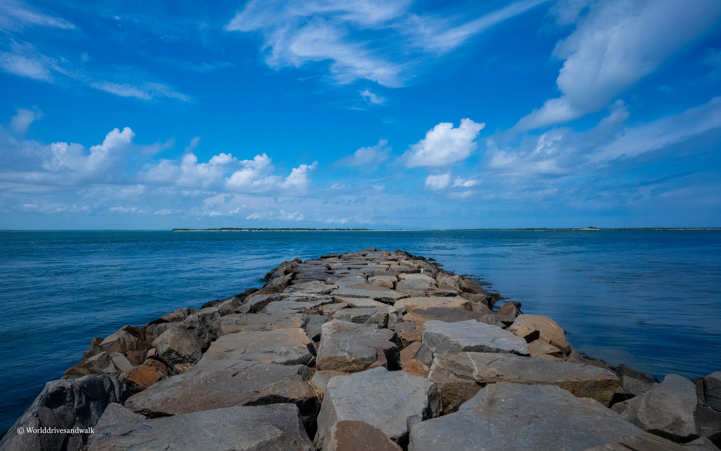 A stone pier extends into the ocean, with blue water and a blue sky with scattered clouds