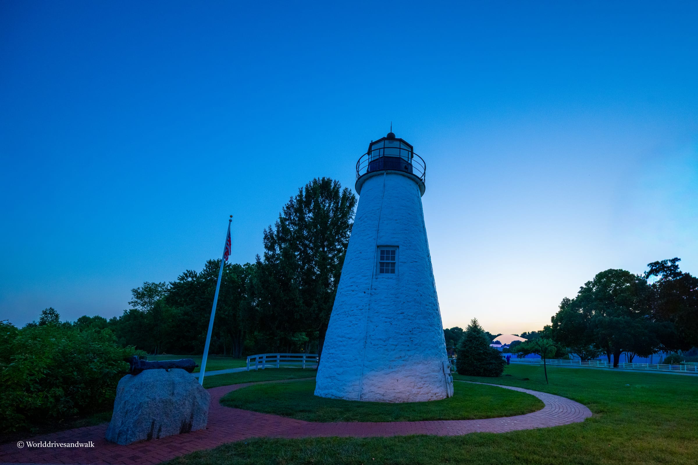Concord Point Lighthouse stands tall against a twilight sky, surrounded by a grassy area with a flagpole and a cannon.