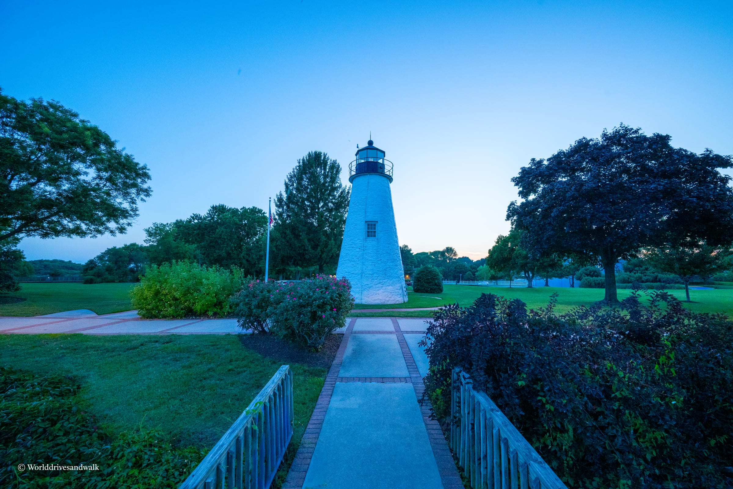 A tall white lighthouse stands in a park with a pathway leading to it, surrounded by green grass, trees, and bushes under a blue sky.