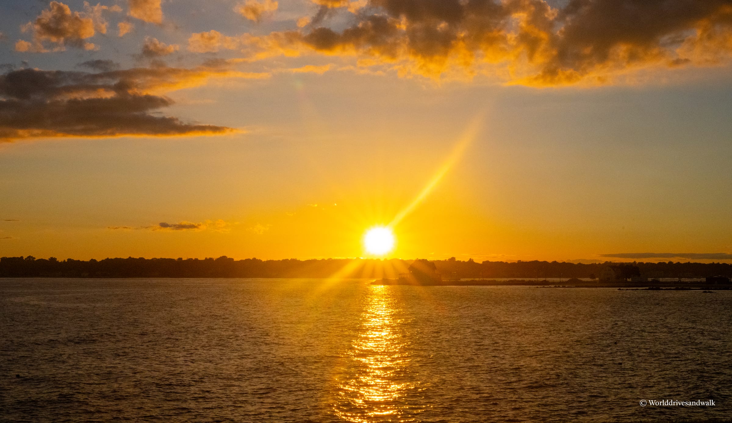 A bright sun sits low on the horizon over calm water, casting golden light and a shimmering reflection. Dark clouds frame the sun above a distant tree line. The sky transitions from deep yellow near the sun to pale gold