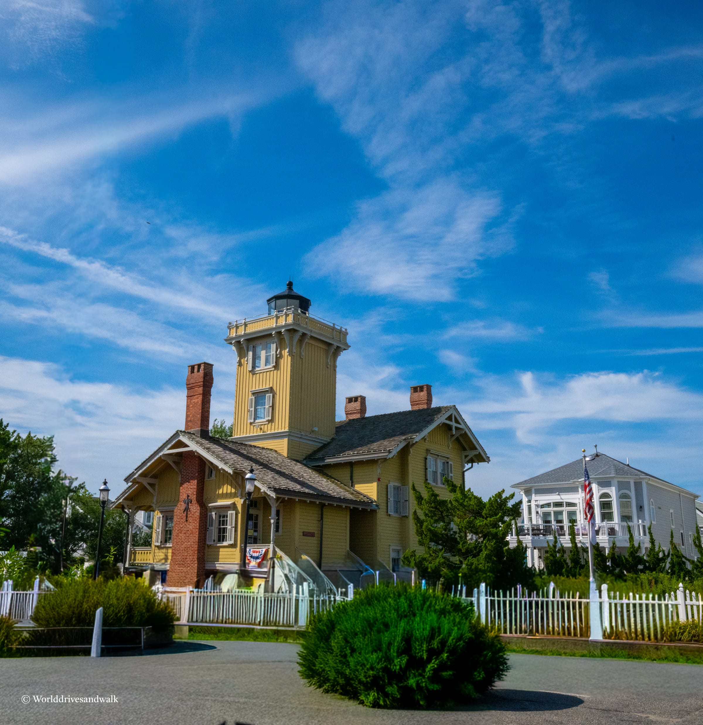 A yellow Victorian lighthouse with a brick keeper's cottage and dark roof stands behind a white picket fence, with a white building visible to the right and blue sky with wispy clouds above.