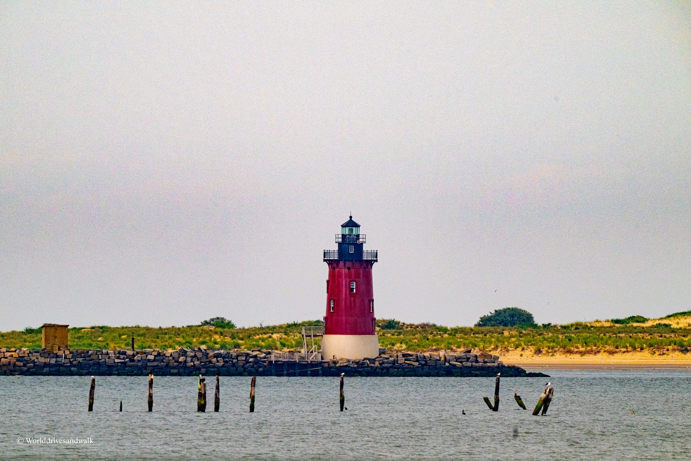 A red and white lighthouse stands on a rocky breakwater with a small building in the background and a sandy beach in the distance.