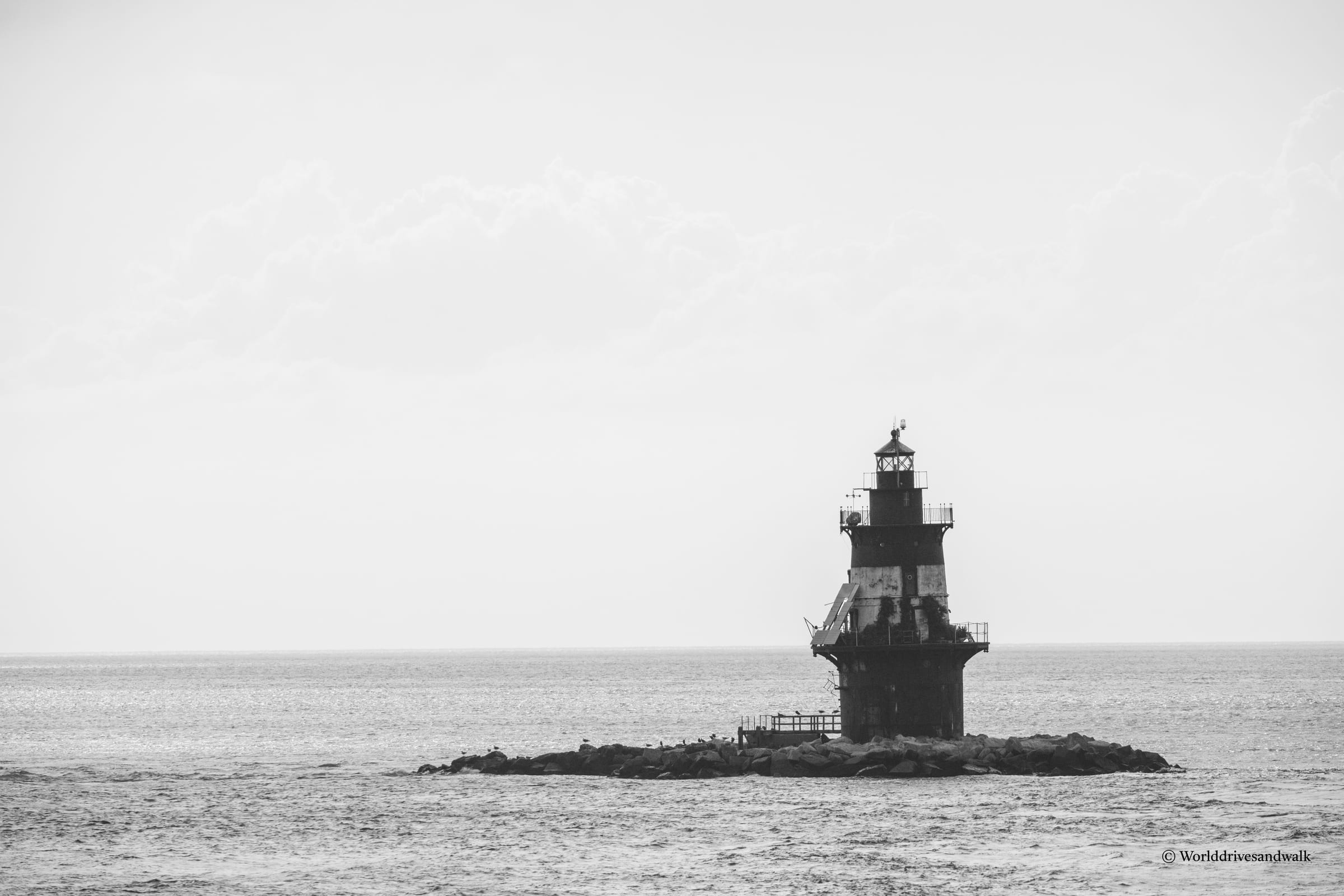 A black and white photograph of Orient Point Lighthouse, a dark cylindrical tower with a lantern room at its top, standing on a small rocky base in calm water. The horizon stretches flat and pale behind it.