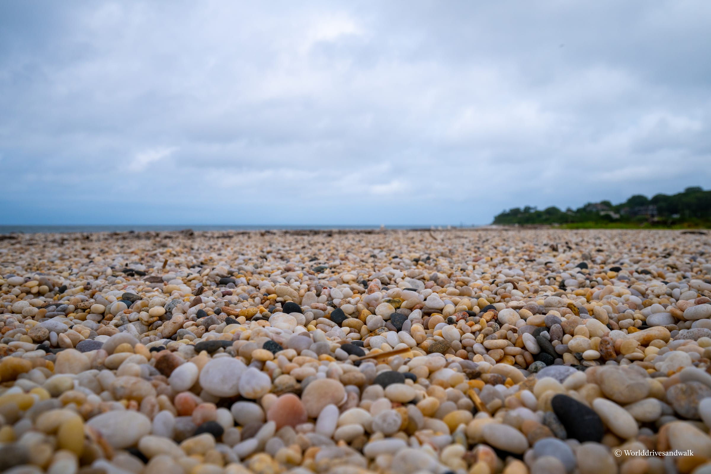 Close-up view of a pebble beach with smooth, rounded stones in cream, tan, and gray tones. The horizon line shows calm water and a cloudy sky, with tree-lined shore visible in the distance.