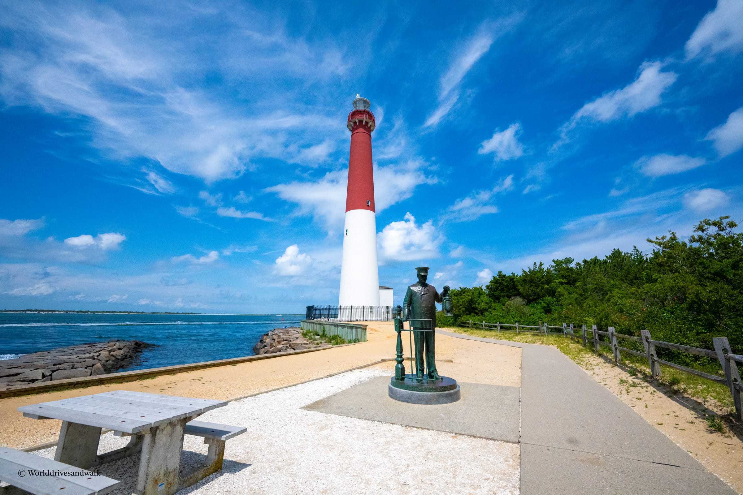 A red-and-white striped lighthouse stands on a sandy point beside the ocean under a blue sky with wispy clouds. A bronze statue of a lighthouse keeper stands on a concrete pad in the foreground, with wooden benches and a
