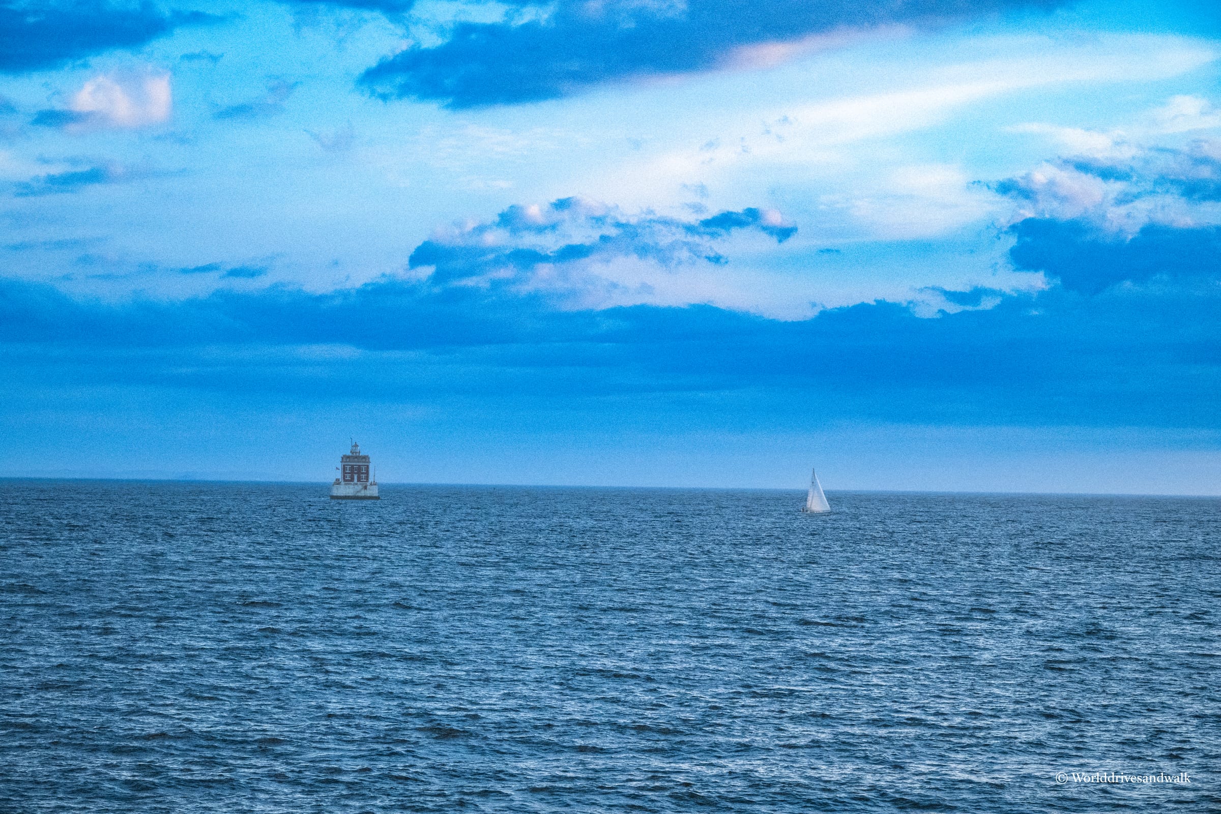 A white sailboat and a stone lighthouse with a lantern room sit on calm blue-green water under a partly cloudy sky.