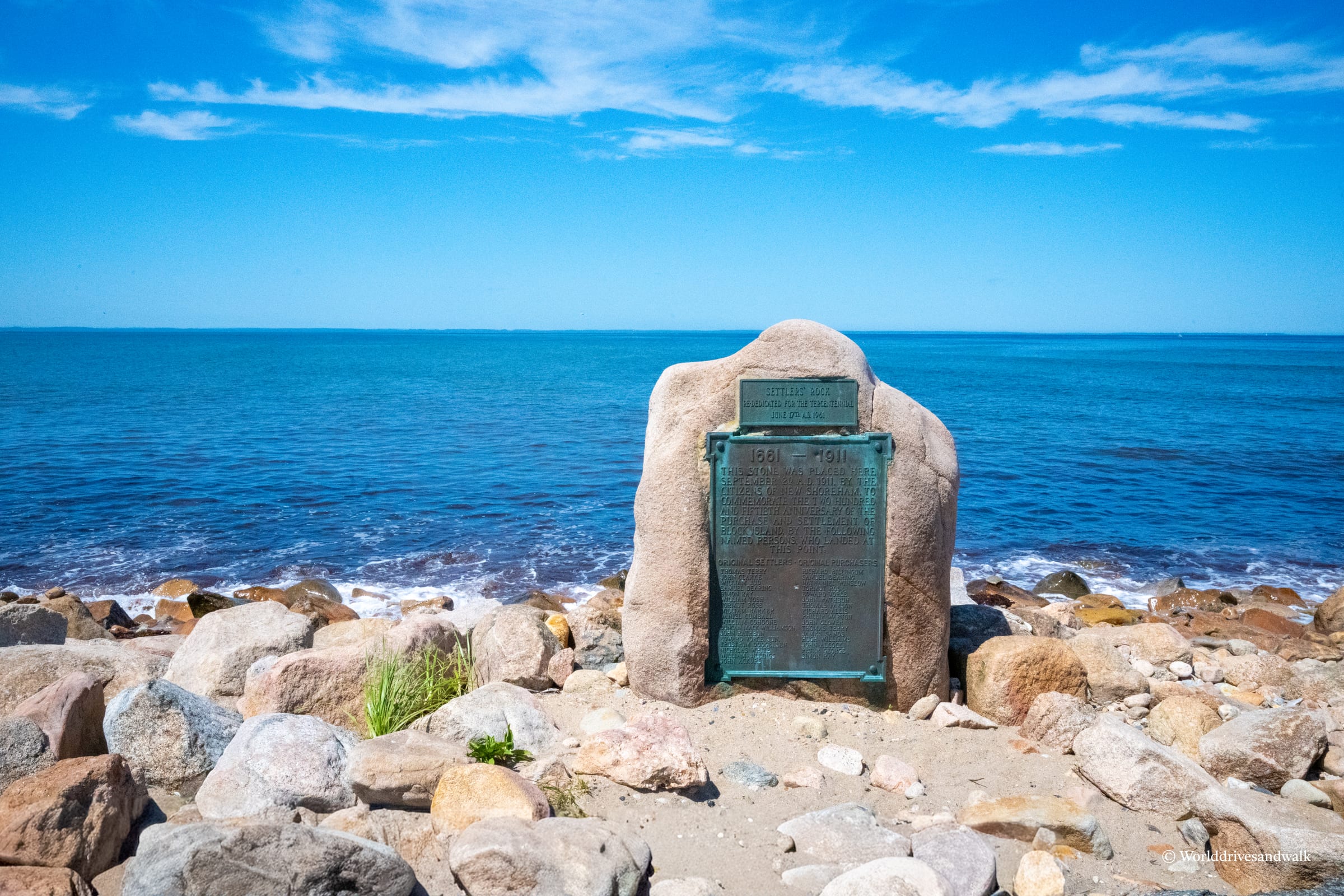 A green plaque mounted on a large rounded boulder sits on a rocky beach with calm blue ocean and sky in the background.