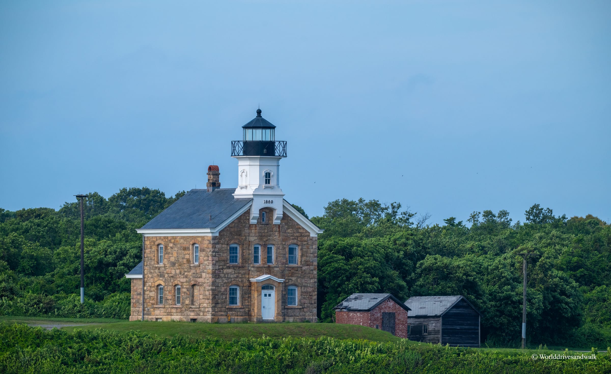A stone lighthouse with white upper tower and black lantern room stands in a grassy field, flanked by two weathered outbuildings, surrounded by dense green trees under clear blue sky.