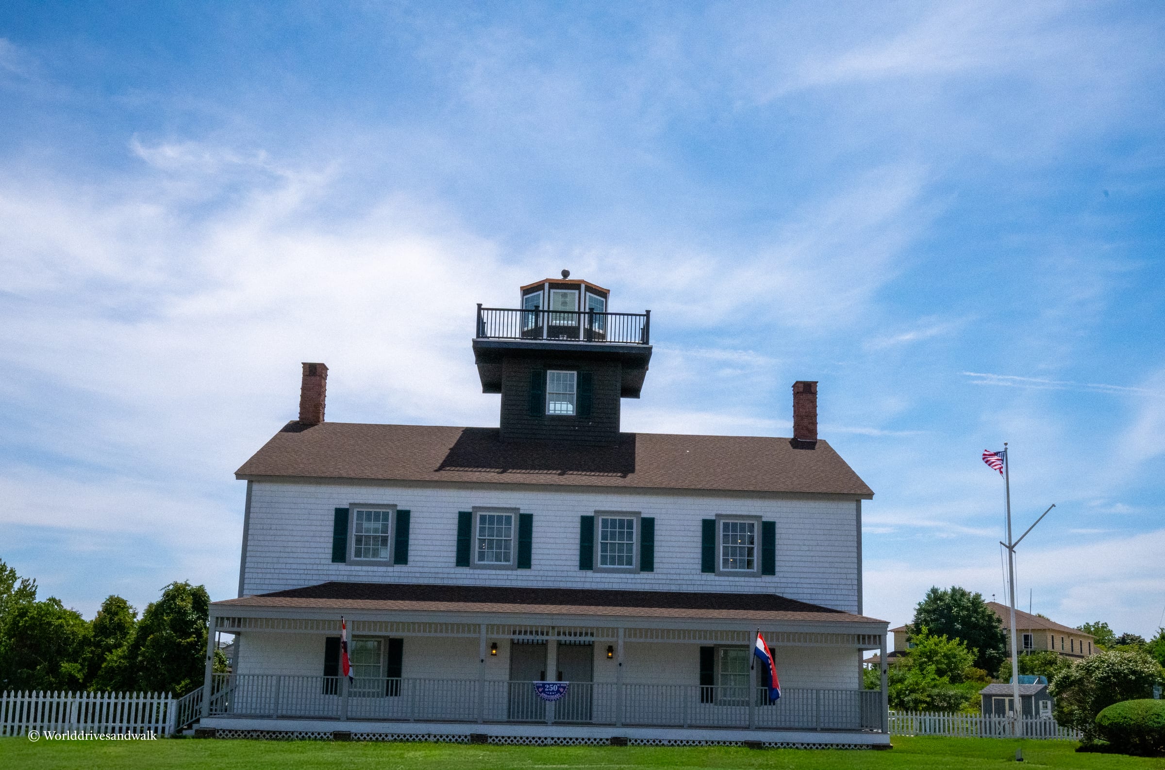 A white two-story keeper's cottage with dark green shutters and two brick chimneys, topped by a square black lighthouse tower with a railed gallery. An American flag flies on the right. The building sits on manicured gre