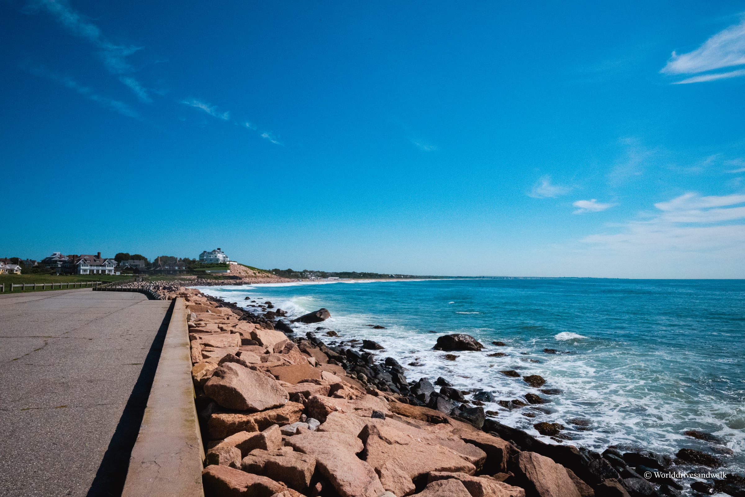 A paved waterfront promenade with a dark railing runs alongside a rocky shoreline. Reddish-brown boulders line the water's edge where waves break against them. Beyond, a sandy beach stretches toward distant houses and gr